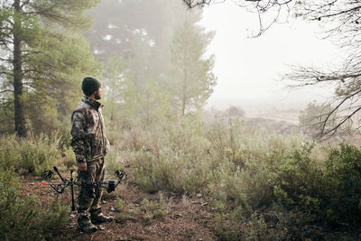 Side view of man in camouflage standing with compound bow in forest and looking away during hunting