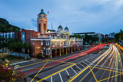 Light trails on city street by buildings against sky