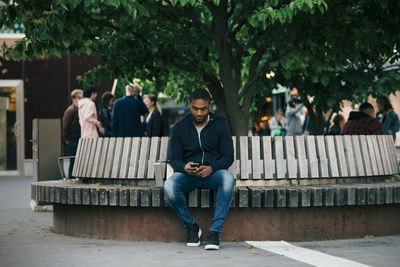 Full length of young man using smart phone while sitting on bench in city