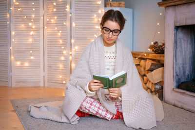  woman with glasses is sitting by fireplace in blanket reading book . cozy atmosphere light garlands