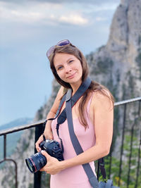Portrait of young woman standing on ai-petri mountain against sky with photo camera 