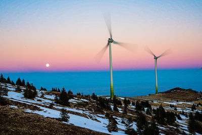 Wind turbines on snow covered landscape against sky during sunset