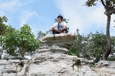 Low angle view of young man sitting on rock against sky