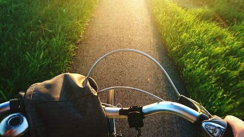 Cropped hand riding bicycle on road amidst grassy field during sunny day