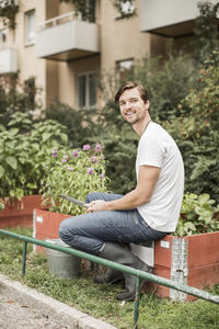 Full length portrait of young man with gardening equipment sitting in garden