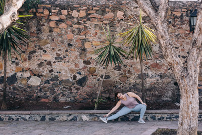 Side view of woman sitting by wall stretching