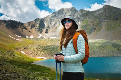 A young caucasian woman with a backpack and in a panama, stands with trekking poles near a mountain