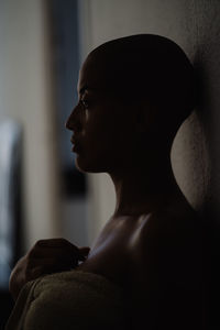 Side view of tranquil african american female with bald hairstyle and in towel leaning on wall at home and looking away