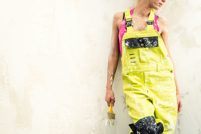 Midsection of young woman in coveralls holding paintbrush against white wall