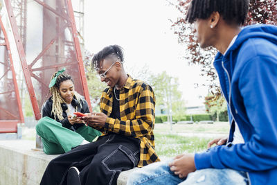 Happy multiracial friends using smart phone sitting on wall