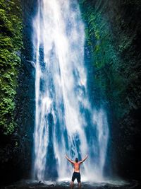 Rear view of shirtless man standing with arms outstretched by waterfall