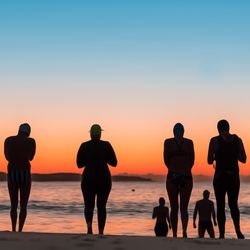 Rear view of people in swimwear while standing at beach against sky during sunset