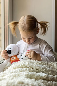 A little girl in white clothes plays with soft toys by the window. 