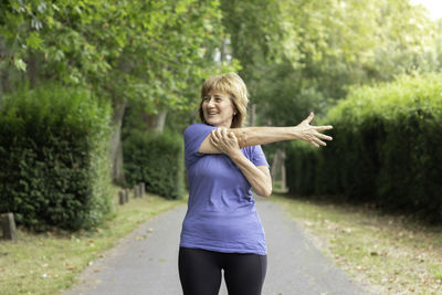 Mature woman in workout outfit smiling to the side streching arm in the green park and trees around