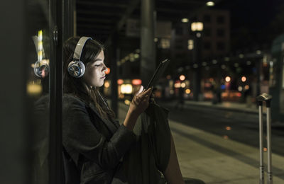 Young woman with headphones waiting at the station by night using tablet