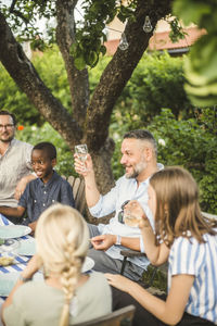 Mature man raising wine glass while sitting with friend and children in backyard party