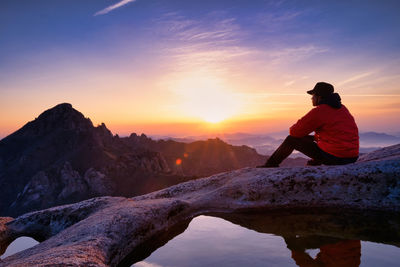 Rear view of man sitting on rock against sky during sunset