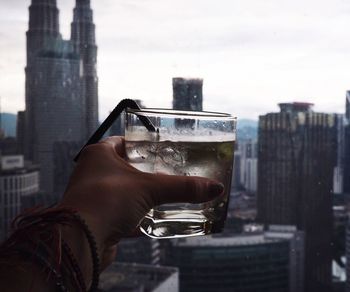 Cropped hand of woman holding drink by window against cityscape
