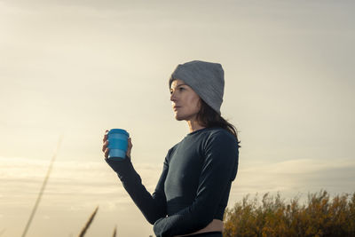 Young woman drinking water while standing against sky during sunset