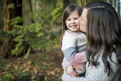 Mother kissing her beautiful cute daughter on the cheek in the forest.