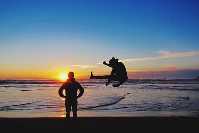 Silhouette man standing by friend jumping at beach during sunset