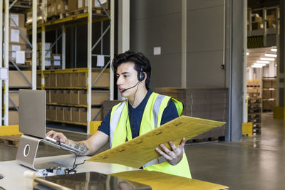 Confident young male customer service representative using laptop while sitting with package at desk in warehouse