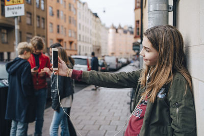 Girl taking selfie through smart phone with friends standing against wall