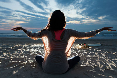 Rear view of young woman with arms outstretched sitting at beach against cloudy sky