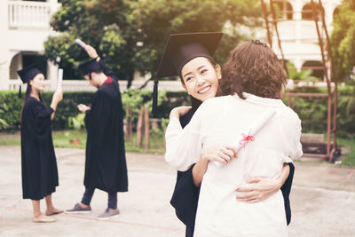 Cheerful woman in graduation gown embracing mother while standing outdoors