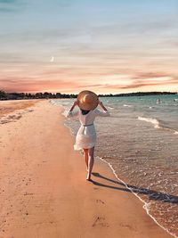 Rear view of woman wearing hat while walking on shore at beach against sky during sunset