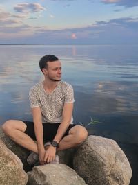 Young man sitting on rock by sea against sky