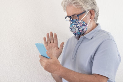 Senior man wearing mask using smart phone standing against wall
