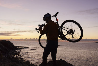 Cyclist carrying bicycle in front of sea at sunrise