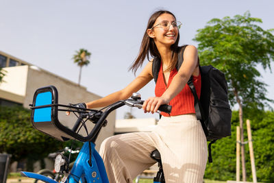 Side view of positive young female with black backpack strolling with bicycle on street of city on sunny summer day