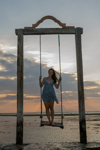 Portrait of happy woman standing on rope swing at beach against sky