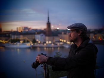 Side view of young man standing by river against city at dusk