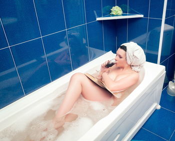 Naked woman with wine reading book while sitting in bathtub