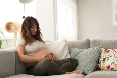 Pregnant mid adult woman sitting on sofa against window at home