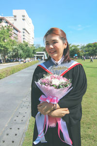 Young woman wearing graduation gown holding bouquet on field during sunny day