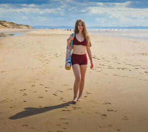 Portrait of smiling young woman walking with exercise mat on beach