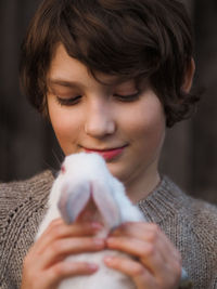 Cute little boy in gray knitted sweater holds a small white rabbit in his arms.