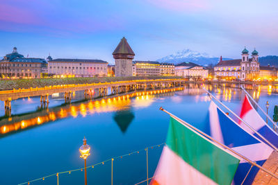 Illuminated buildings by river against sky at dusk