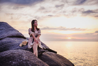 Woman sitting on rock by sea against sky during sunset