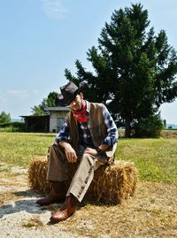 Full length of cowboy sitting on hay at field against sky