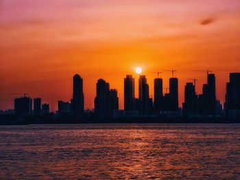 Silhouette buildings by sea against sky during sunset