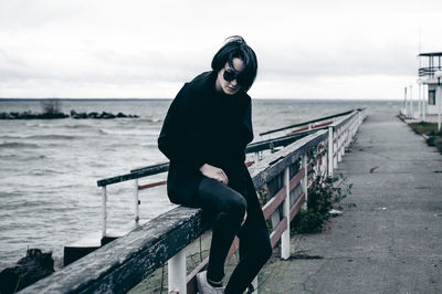 Woman sitting on railing by sea against sky