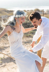 Bride sitting on rope while man helping outdoors