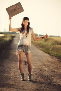 Full length portrait of woman hitchhiking while holding text on road