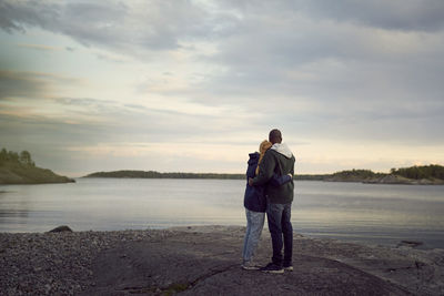 Rear view of couple standing at beach while looking at lake against sky during sunset