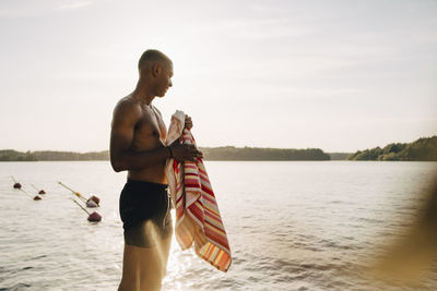 Smiling shirtless wet man with towel looking at lake in summer